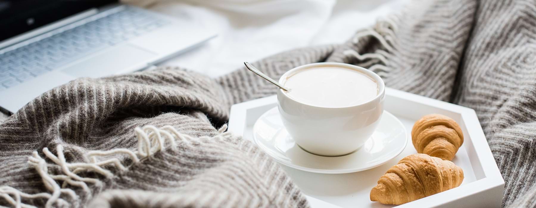 lifestyle image of a mug of coffee, a couple of croissants and a grey blanket